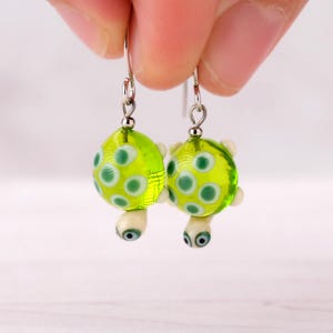 Puede incluir: Pendientes hechos a mano con diseño de tortuga. Cada pendiente tiene un cuerpo de vidrio redondo verde brillante con lunares blancos y verdes. La cabeza y las patas de la tortuga son blancas con ojos azules, y los pendientes cuelgan de ganchos plateados.