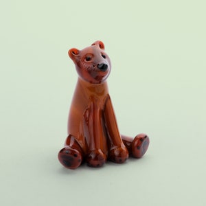 Peut inclure: Une petite figurine d'ours en verre brun avec des yeux noirs et des empreintes de pattes. L'ours est assis la tête penchée vers le haut.