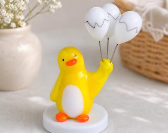 May include: A yellow penguin ornament with a white belly, orange feet, and a small orange beak. The penguin holds three white balloons with black zigzag lines. The ornament sits on a white circular base.