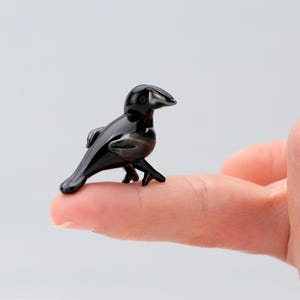 May include: A small black glass crow figurine with a detailed design. The crow is perched on a finger.