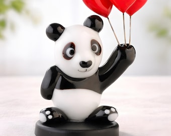 May include: A ceramic panda figurine in black and white, holding three red heart-shaped balloons. The panda is sat on a black base, with its arm raised. The figurine has a glossy finish.