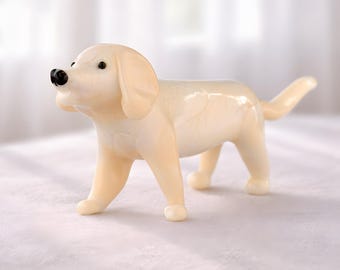 May include: A cream-coloured dog figurine with a glossy finish. The dog has a black nose and small, dark eyes. It is standing on a white surface, with a soft, blurred background.