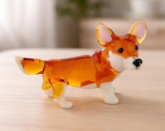 May include: A glass corgi ornament in a standing pose. The dog has a translucent orange body with white paws and chest. The ears are pink-lined, and the nose is black. The ornament is on a light-coloured surface.