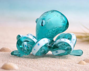 May include: A small glass octopus figurine in a translucent teal colour. The octopus features a rounded head with embedded silver-toned metallic flakes and eight curled tentacles decorated with small, evenly spaced white polka dot patterns.