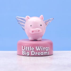 Może przedstawiać: Różowa szklana figurka świnki z przezroczystymi szklanymi skrzydłami, stojąca na różowej podstawie z napisem "Little Wings Big Dreams".