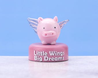May include: A pink glass pig figurine with clear glass wings, standing on a pink base with the words "Little Wings Big Dreams".