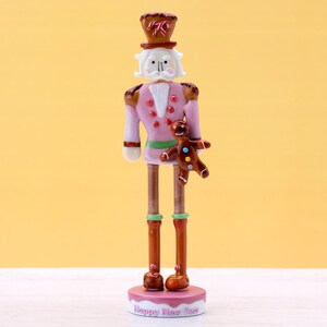 Puede incluir: Una figurita de cascanueces de jengibre rosa y marrón que sostiene un hombre de jengibre. El cascanueces lleva una faja verde y un sombrero marrón con un lazo rojo. La base de la figurita dice "Happy New Year".