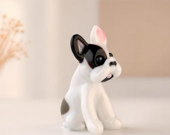 May include: A glossy white and black French Bulldog ornament. The dog has black ears, a black patch on its back, and pink detailing on its ears and mouth. The ornament is sitting on a white surface.