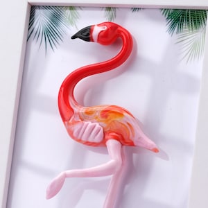 Może przedstawiać: Szklana rzeźba flaminga w kolorze różowym i pomarańczowym w białej ramie. Flamingo stoi w pozycji stojącej z głową odwróconą w bok.