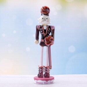 Può includere: Una statuetta di schiaccianoci in vetro con un design dipinto di rosa e marrone. La statuetta tiene un bastone di zucchero rosa e marrone. La base della statuetta è rosa e dice "Happy Nutcracker Day".