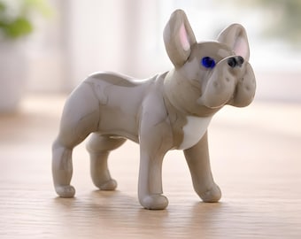 May include: A grey ceramic French Bulldog ornament with blue eyes and a white patch on its chest. The dog is posed on all fours, with its ears erect. This is a decorative item.