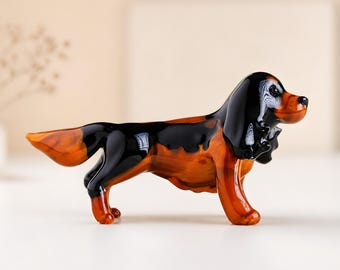 May include: A glass dog figurine with a glossy finish. The dog is depicted in a standing pose, with a black head and back, and a reddish-brown body. This decorative figurine would make a charming addition to any home.