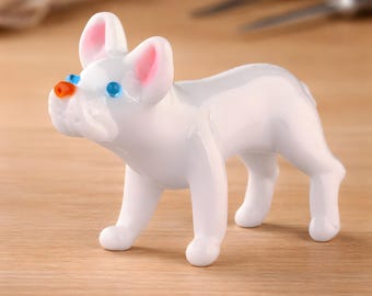 May include: A white French Bulldog figurine with pink-lined ears, blue eyes, and an orange nose. The dog is posed on all fours, with a glossy finish. The figurine is set on a light-coloured wooden surface.