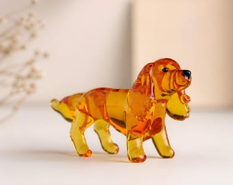 May include: A decorative glass dog figurine in amber. The dog has long, floppy ears and a black nose. The figurine is posed standing. The translucent glass allows light to pass through. A small ornament, likely for display.