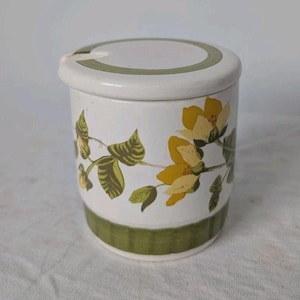 May include: A white ceramic canister with a lid featuring a green band around the top and bottom. The body is decorated with a floral pattern of yellow flowers and green leaves. The lid includes a small notch for a spoon or utensil to rest inside.