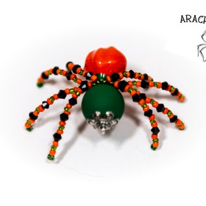 May include: A beaded spider with an orange head, green body, and black and orange legs. The spider is sitting on a white surface. The text "ARACH-N-SPIRED" is in the upper right corner of the image.