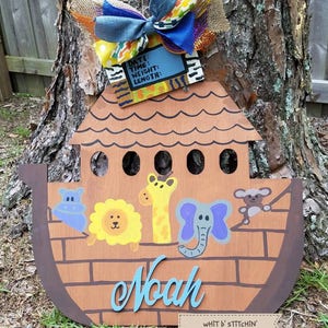 May include: A wooden Noah's Ark door hanger painted in brown, blue, yellow, and purple with a blue and white name "Noah" and the text "WHITT B' STITCHIN'" on a brown tag. The ark has a brown roof with a brown bow and a blue bow with colorful ribbons on top.