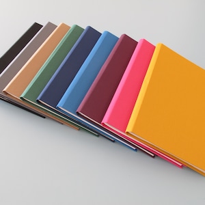 Puede incluir: Una pila de diez cuadernos con cubiertas de diferentes colores. Las cubiertas son negras, grises, marrones, verdes, azul marino, azules, burdeos, rosas y amarillas.