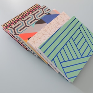 Op de afbeelding: Een stapel van vijf notitieboekjes met verschillende geometrische patronen op de omslagen. De omslagen zijn in tinten blauw, groen, oranje, roze en zwart.
