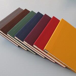 Könnte beinhalten: Ein Stapel von acht Notizbüchern mit verschiedenen farbigen Umschlägen. Die Umschläge sind grau, braun, grün, blau, burgunderrot, rot und gelb.