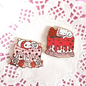 May include: Two enamel pins featuring adorable animal characters sleeping on top of pink and red cakes. The pink cake has a panda bear sleeping on top and the red cake has a white cat sleeping on top. Both cakes have white frosting and red hearts.