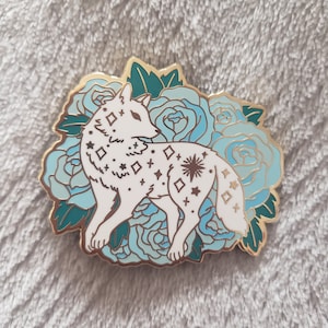 Puede incluir: Un pin de esmalte blanco de un lobo con un patrón estrellado, rodeado de flores de esmalte azul y verde.