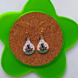 Puede incluir: Un par de pendientes de aro dorados con un diseño de bombilla. Cada bombilla presenta un gato negro en un bote con un remo, rodeado de nubes blancas y un mar azul.