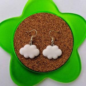 May include: A pair of white cloud-shaped earrings with silver hooks. The earrings are displayed on a green star-shaped cork coaster.