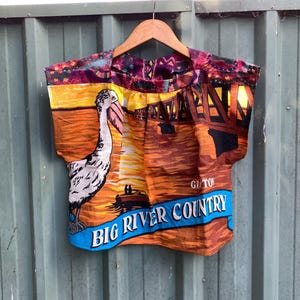 May include: A sleeveless top with a vibrant print depicting a pelican, a bridge, and the words "Big River Country." The bottom has a blue band with white text. The background shows a sunset with orange and yellow tones.