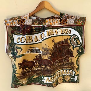 May include: A sleeveless top featuring a vintage-style graphic of a Cobb & Co. stagecoach with the text "COBB & CO. 1854-1924", "Greetings from TOOWOOMBA THE GARDEN CITY", and "AUSTRALIA". The neckline has a patchwork border with floral and paisley patterns.