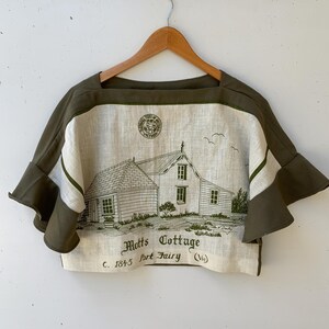 May include: Olive green and cream-coloured crop top with a square neckline and flared sleeves. The front features a detailed illustration of a cottage with the text "Motts Cottage C. 1845 Port Fairy (Vic)". The top is hanging on a wooden hanger.