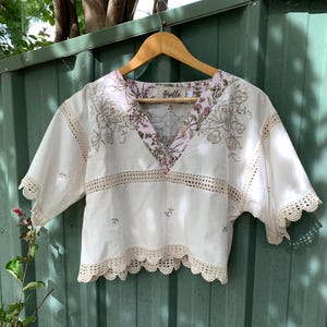 Könnte beinhalten: Weißes Crop-Top mit V-Ausschnitt und kurzen Ärmeln. Das Oberteil hat Blumenstickereien, Spitzensaum und einen rosa geblümten Ausschnitt. Der Markenname "Grelle" ist auf einem Etikett zu sehen. Das Oberteil hängt an einem Holzbügel.