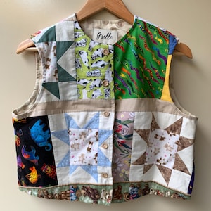 May include: A patchwork vest with a variety of fabric patterns, including stars, bears, and abstract shapes. The vest has a button-up front and a wooden hanger. The label reads "giselle".