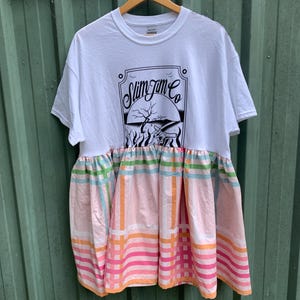 Könnte beinhalten: Weißes T-Shirt mit einer schwarz-weißen Grafik eines Logos mit der Aufschrift "Slim Jim Co.". Das T-Shirt hat einen rosa-grünen karierten Rock, der am unteren Rand befestigt ist.