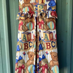 May include: A baby changing pad cover with a repeating pattern of teddy bears, dolls, ABC blocks, and colorful balls. The top edge features a ruffle. The color palette includes brown, red, blue, yellow, and white.