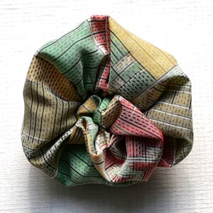 May include: A knotted fabric item with a plaid design in green, yellow, red, and tan. The fabric is gathered and tied, forming a textured, rounded shape. The background is a plain white surface.