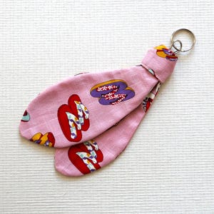 Op de afbeelding: Een roze stoffen sleutelhanger met een patroon van Japanse geta-sandalen in rood, paars en geel. De sleutelhanger heeft een zilveren metalen ring om aan sleutels of tassen te bevestigen. Geta-sandalen zijn traditioneel Japans schoeisel.
