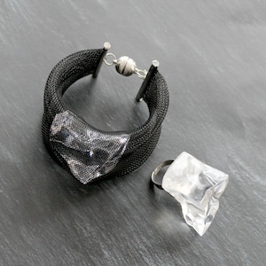 Op de afbeelding: Een zwarte mesh armband met een zilveren sluiting en een transparante plastic ring met een geometrisch ontwerp.