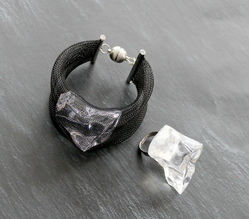 Op de afbeelding: Een zwarte mesh armband met een zilveren sluiting en een transparante plastic ring met een geometrisch ontwerp.