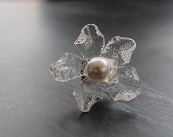 Op de afbeelding: Een heldere, bloemvormige ring met een zilveren middenstuk. De bloemblaadjes hebben een textuur, ijzig uiterlijk. De ring staat tegen een donkere achtergrond, wat het delicate ontwerp benadrukt.