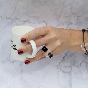 May include: A white ceramic mug with black text that reads "Stay wild, stay outside". A hand is holding the mug, wearing a black ring with a clear glass orb and a black bracelet with clear glass orbs.