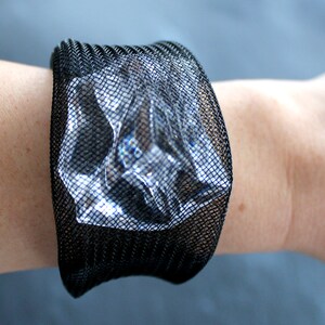 Op de afbeelding: Een zwarte mesh armband met een transparante plastic insert. De armband zit om een pols.