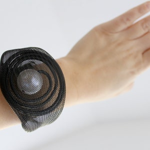 Könnte beinhalten: Ein schwarzes Mesh-Armband mit einem runden Design. Das Armband besteht aus mehreren Schichten Mesh, die einen Spiraleffekt erzeugen. Die Mitte des Armbands ist ein klares, rundes Stück Material.