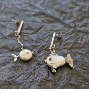 Può includere: Un paio di orecchini a forma di pesce in argento. Ogni orecchino presenta un corpo di perla, un piccolo occhio di pietra preziosa e una pinna caudale argentata. Uno pende da una catena, l'altro è attaccato direttamente a un perno.