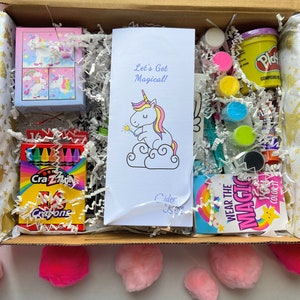 Op de afbeelding: Een knutselset met eenhoorn-thema in een kartonnen doos. De set bevat kleurpotloden, klei, verf, een kleurplaat en een eenhoorn-sticker. De doos is gevuld met wit gescheurd papier. De tekst op de kleurplaat luidt "Let's Get Magical!" en "Cider Rose".