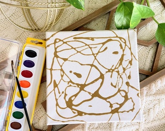 May include: A white canvas with a gold abstract design created using a paint pouring technique. The canvas is on a wooden tray with a white knitted surface underneath. A yellow watercolor paint tray with a brush is also visible.
