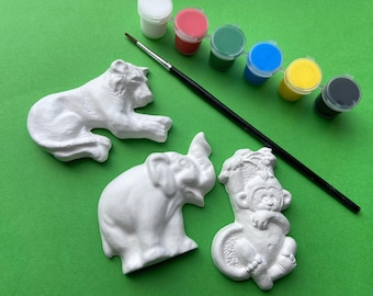 Puede incluir: Un conjunto de cuatro figuras de animales de cerámica blanca, que incluyen un león, un elefante, un mono y un pájaro, con seis botes de pintura de varios colores, incluyendo blanco, rojo, verde, azul, amarillo y negro.