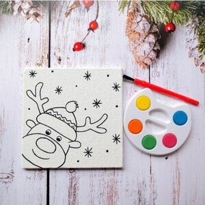 May include: A white canvas with a black and white line drawing of a reindeer wearing a hat. The canvas is next to a white paint palette with six colorful paint wells.