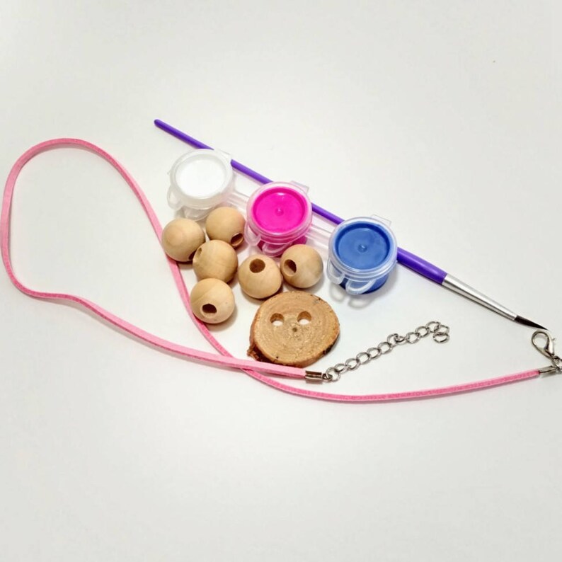 May include: A craft kit for making a necklace with wooden beads, a chain, a pink cord, and three small paint pots: white, pink, and blue. A paintbrush is included.