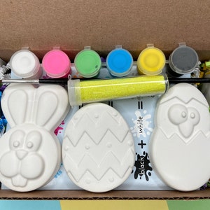 May include: Easter craft kit with three white ceramic figurines: a bunny, an egg, and a chick. The kit includes six small bottles of paint in various colors, a bottle of glitter, and a paintbrush. The kit also includes a sheet of paper with instructions and a black and white color chart.